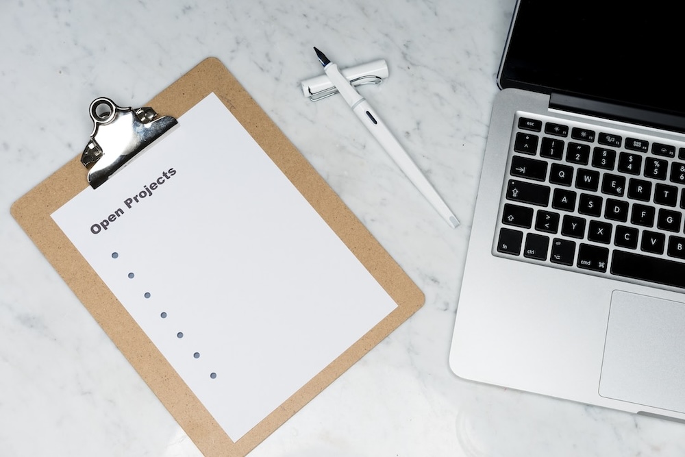 A clipboard with a blank list titled Open Projects sits by a laptop keyboard, a pen between
        them